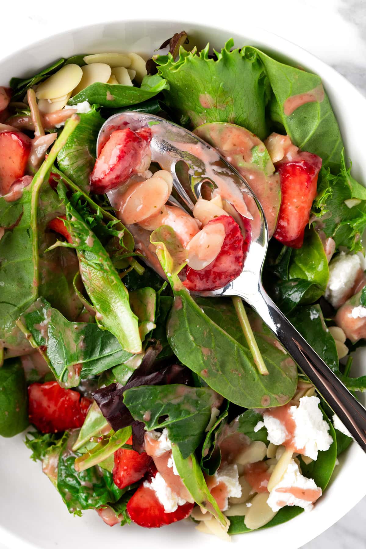 A large metal serving spoon in the bowl of strawberry almond and spinach salad that has been mixed.