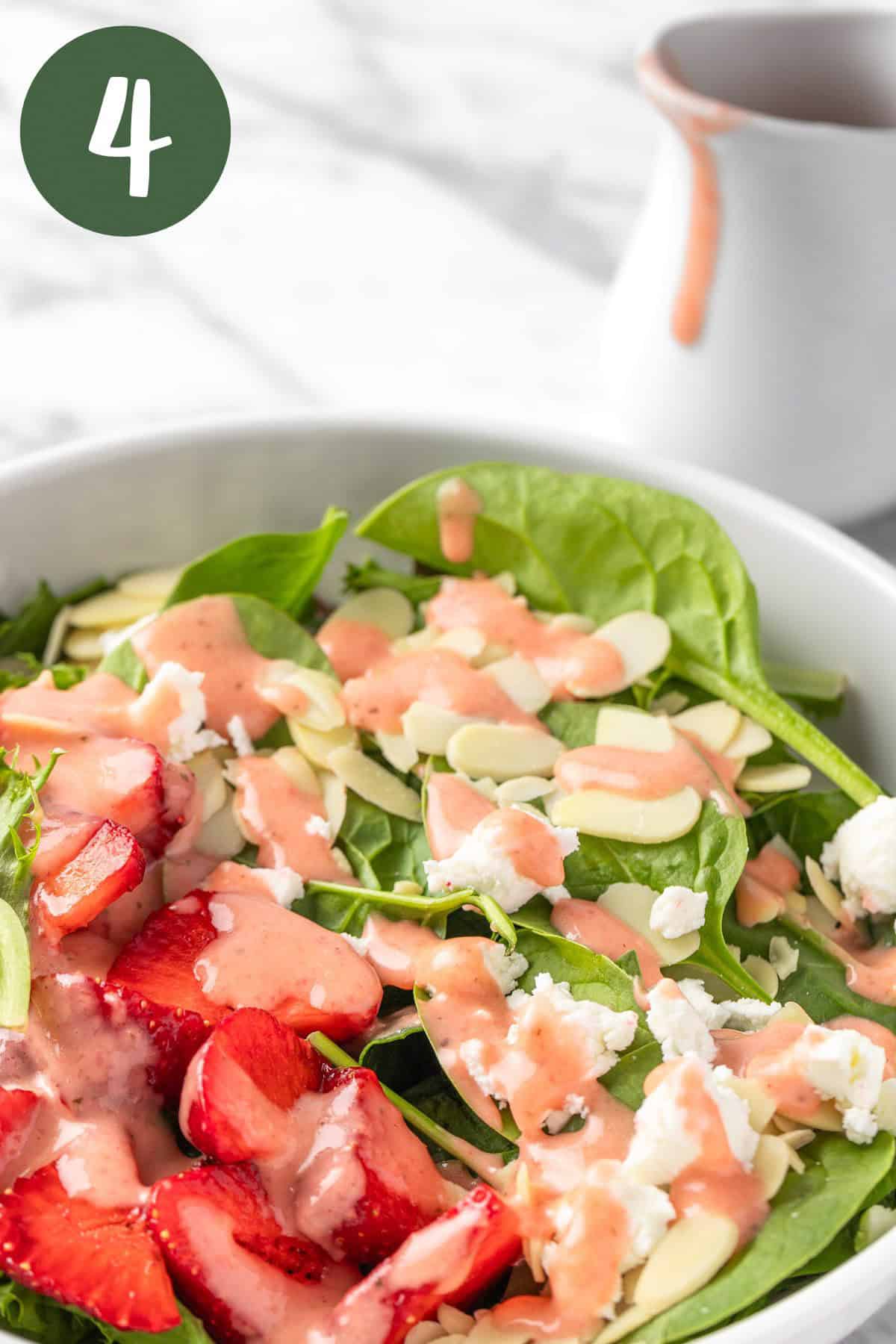 A bowl of spinach strawberry almond salad with a white container of salad dressing in the background.
