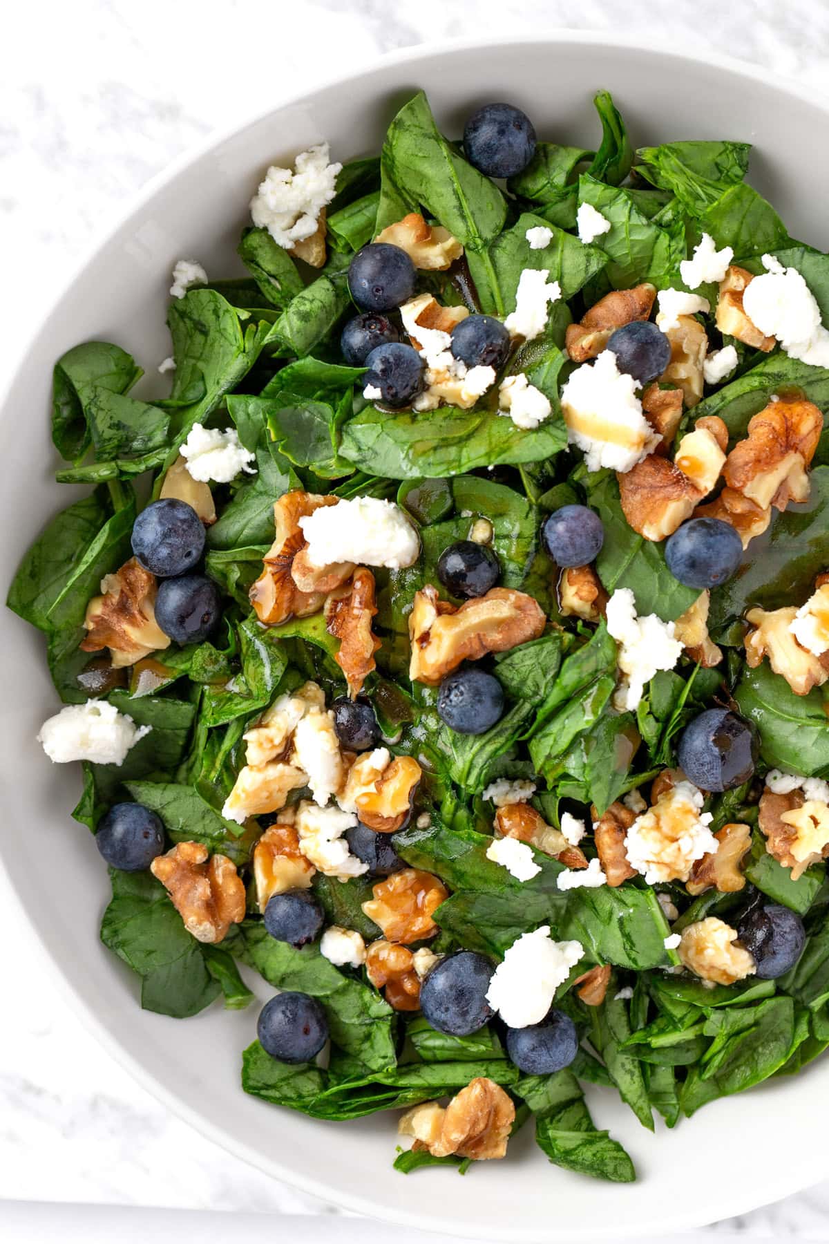 Blueberry goat cheese salad with spinach and toasted walnuts in a white bowl, topped with a maple dressing.