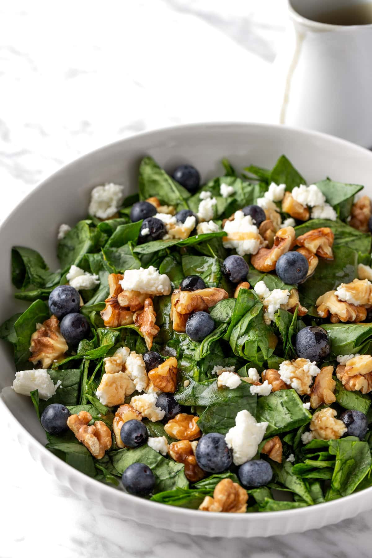 Blueberry goat cheese salad with spinach and toasted walnuts in a white bowl, topped with a maple dressing.
