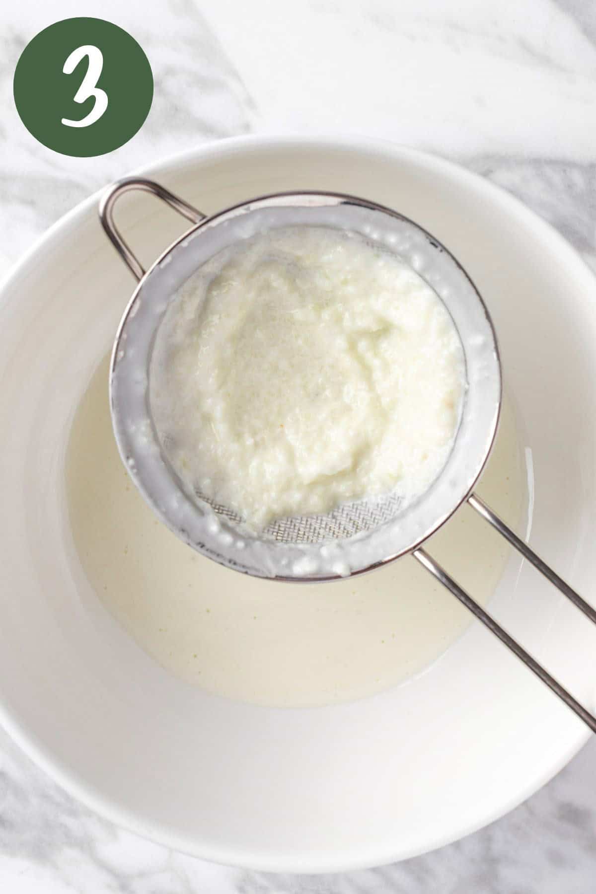 A strainer straining the tzatziki dressing into a white bowl.