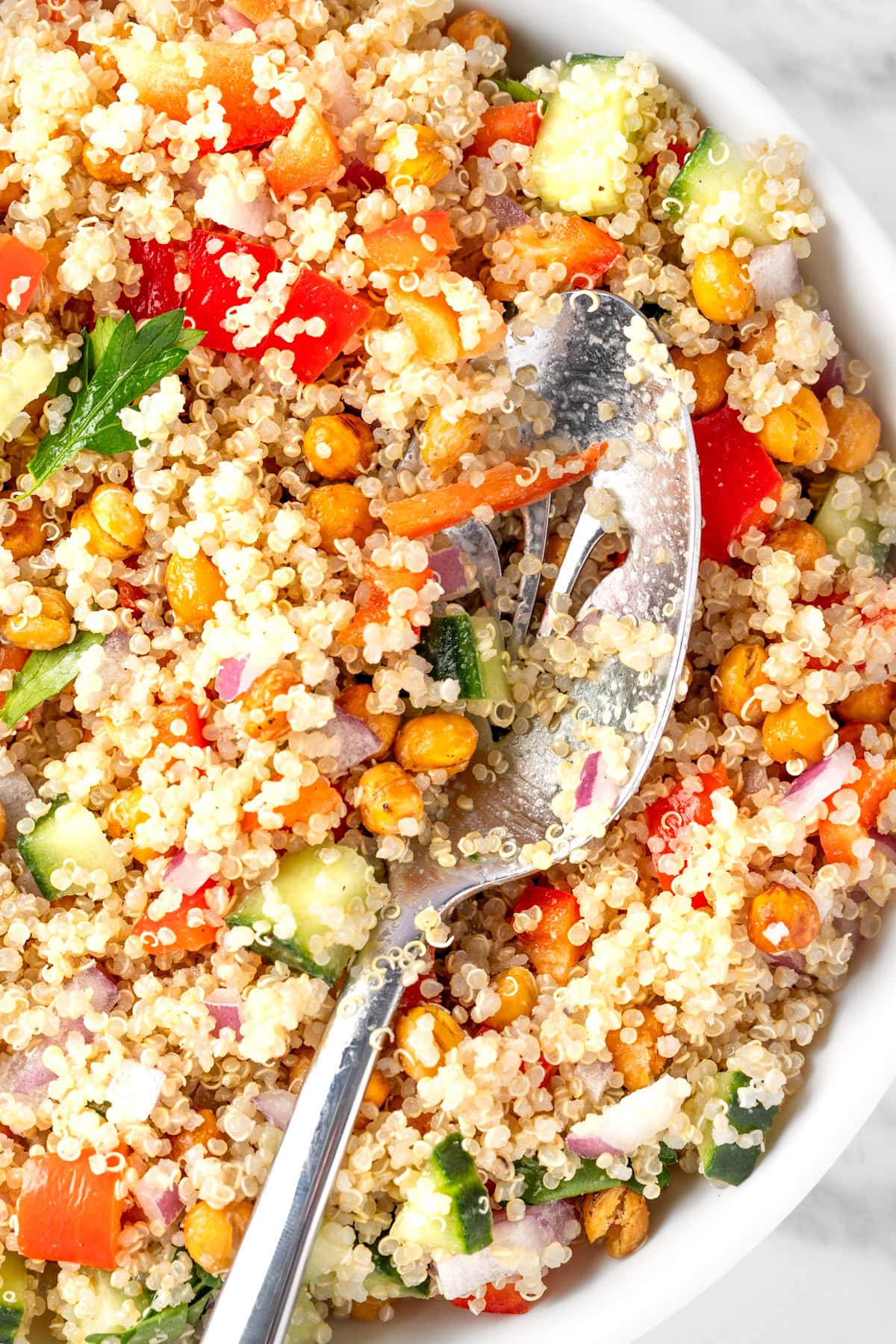 A bowl of quinoa chickpea salad with red bell peppers, cucumber, red onions and parsley, served with a large metal salad spoon.