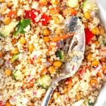 A bowl of quinoa chickpea salad with red bell peppers, cucumber, red onions and parsley, served with a large metal salad spoon.