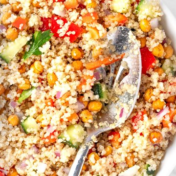 A bowl of quinoa chickpea salad with red bell peppers, cucumber, red onions and parsley, served with a large metal salad spoon.