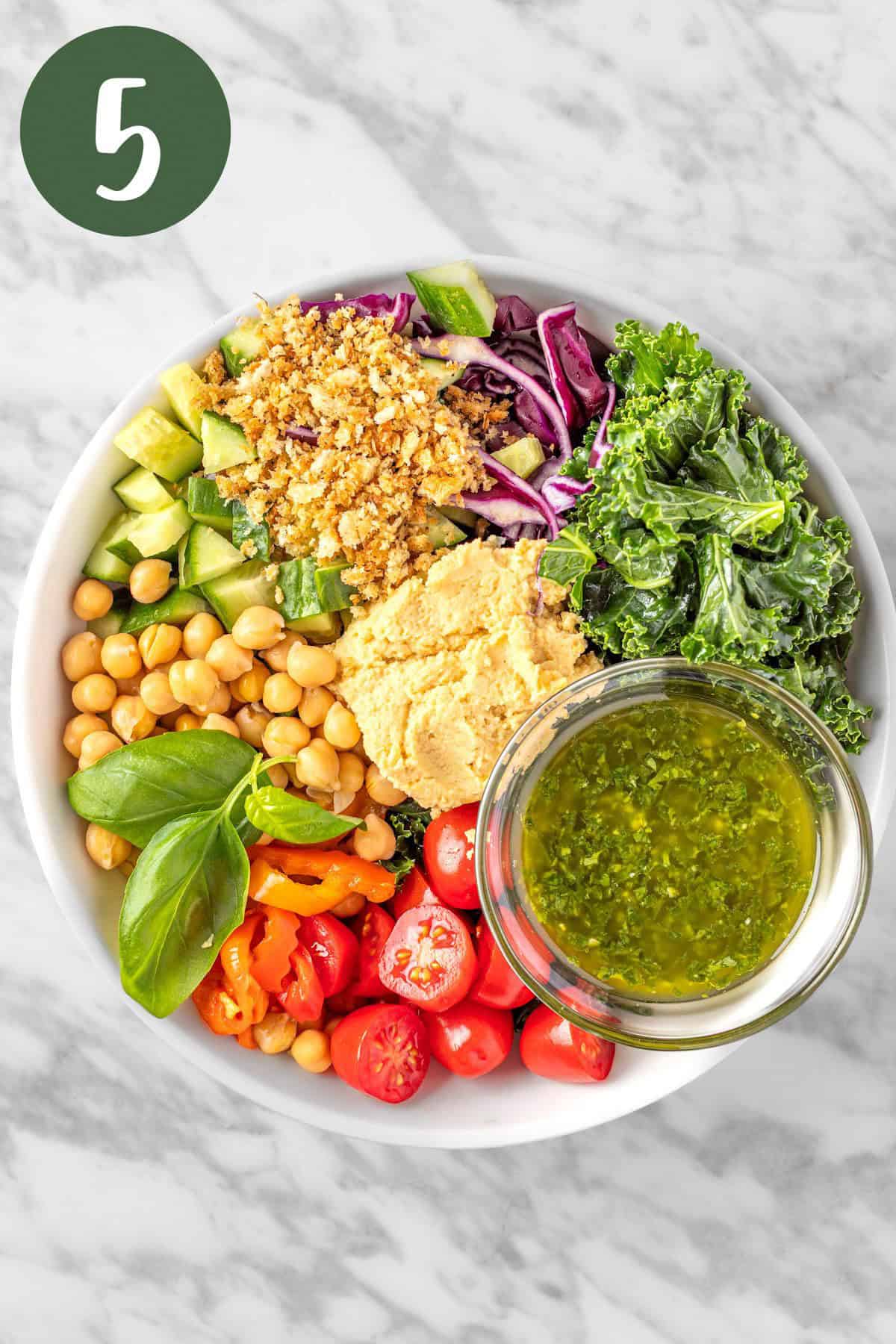 A process photo showing all the salad ingredients added together in a large salad bowl served with a dollop of hummus and pesto vinaigrette.