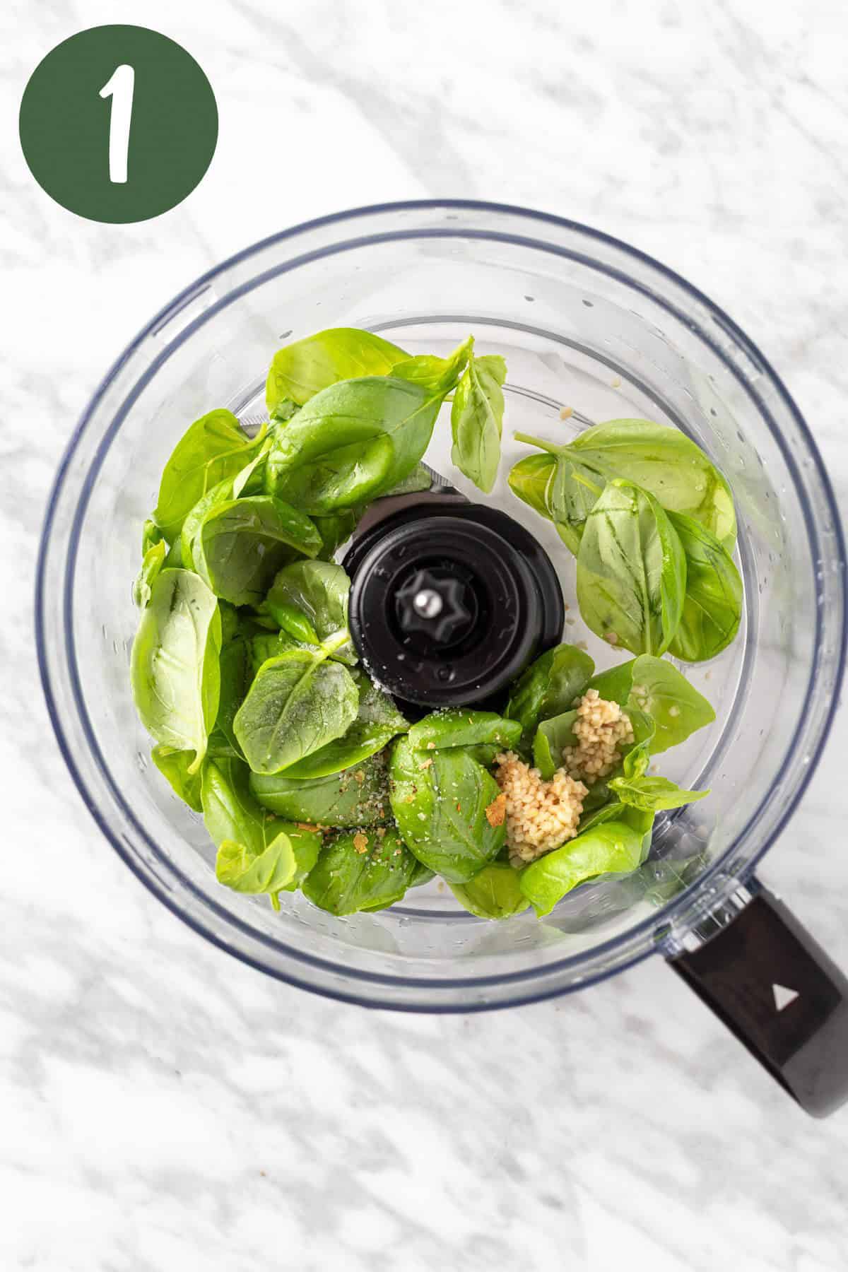A process photo of the basil leaves, seasoning and garlic in a food processor, ready to be blended.