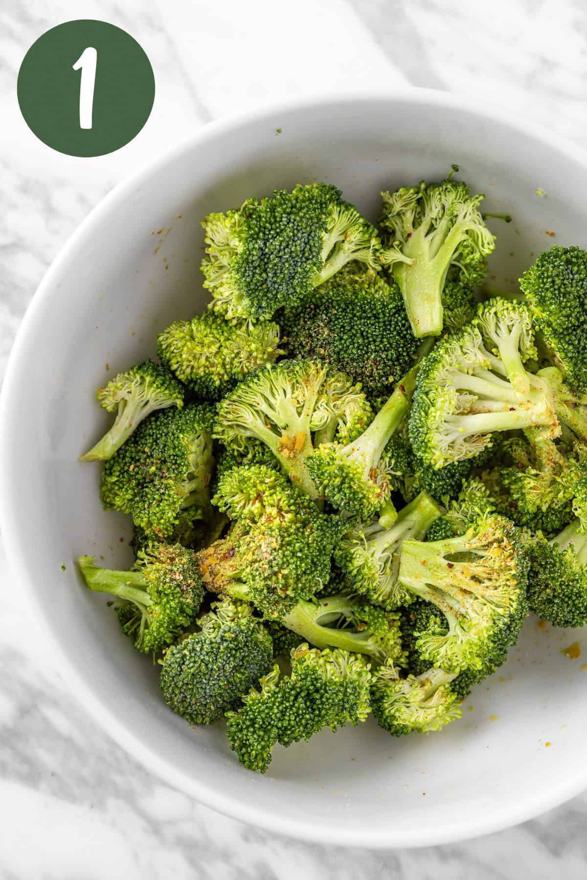 A process photo of the raw broccoli florets in a white bowl, seasoning with olive oil and umami seasoning.